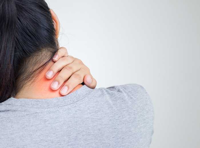 A person in a grey shirt holds their neck with one hand, appearing to soothe a painful, red area on the neck; background is plain and light-coloured.