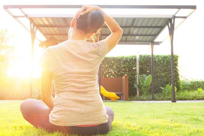 A person sits on grass in a yard, stretching by holding their head with one hand, facing a metal canopy structure, with sunlight streaming from the side.