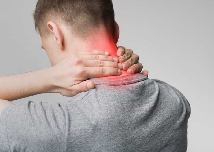 Person holding the back of their neck in apparent pain, highlighted by a red glow, while wearing a grey shirt in a neutral, greyish background.