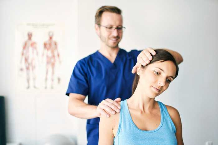 A therapist in blue scrubs adjusts a woman's neck and shoulder in a clinic with anatomical posters on the wall behind them.